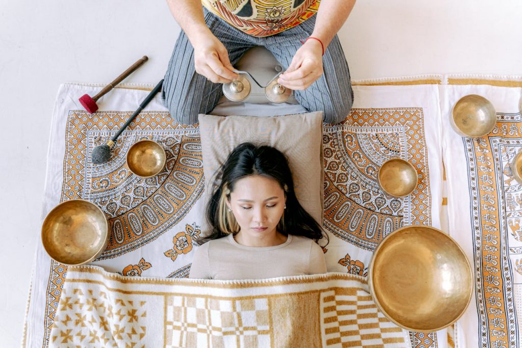 A woman lying on her back with head relaxed on pillow, eyes closed, and Tibetan Sing Bowls around her.