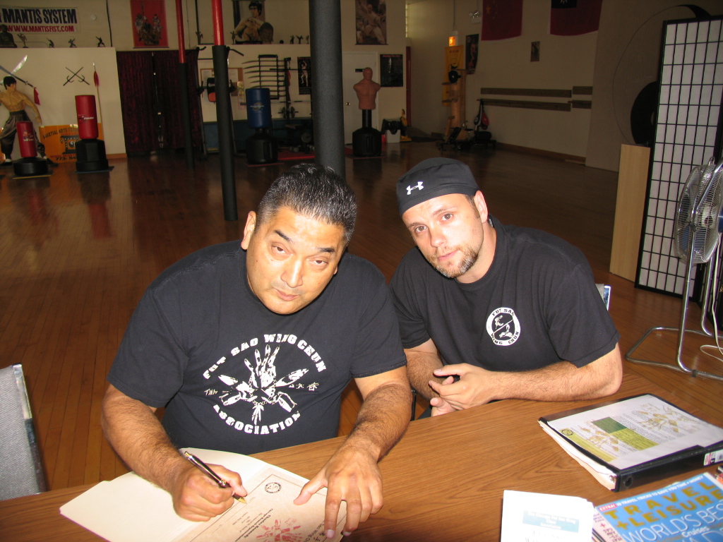 Sigung James Cama and Chuck Kennedy sitting down together at original Fut Sao Wing Chun Association School, both looking at camera while Sigung James Cama signs something.